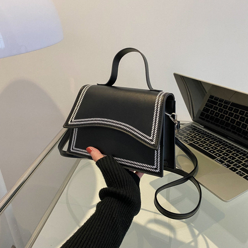 Black handbag with white stitching held by a person next to a laptop on a glass surface.
